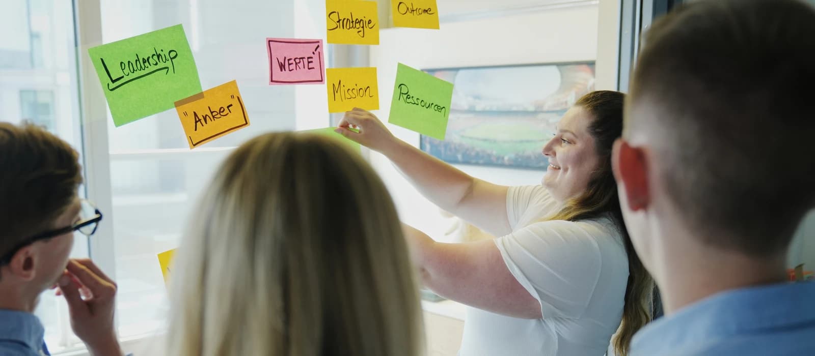 Workshop für Auszubildende: Trainerin hängt Haftnotizen mit Begriffen wie Leadership, Werte und Mission an ein Fenster, die Gruppe schaut zu