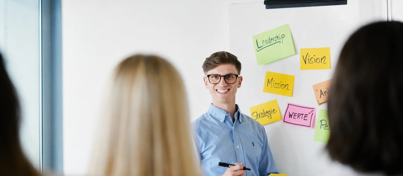 Systemisches Entwicklungsprogramm für Führungskräfte: lächelnder Mann am Flipchart mit Haftnotizen zu Leadership, Vision, Mission und Werte, Teilnehmende im Vordergrund