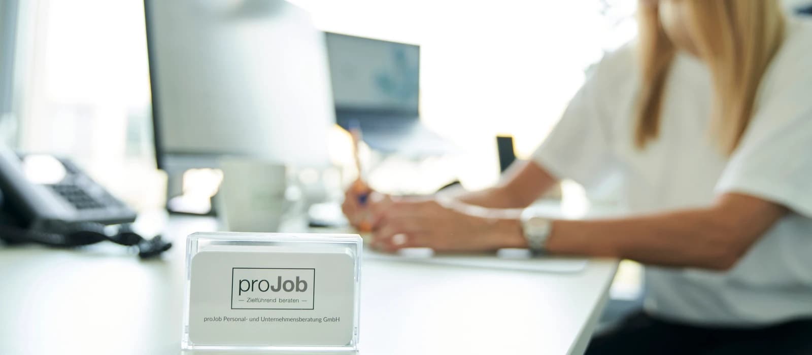 Versicherungsspezialist für Transportversicherungen gewinnen: Visitenkarten-Aufsteller von proJob auf einem Schreibtisch, im Hintergrund schreibt eine Frau